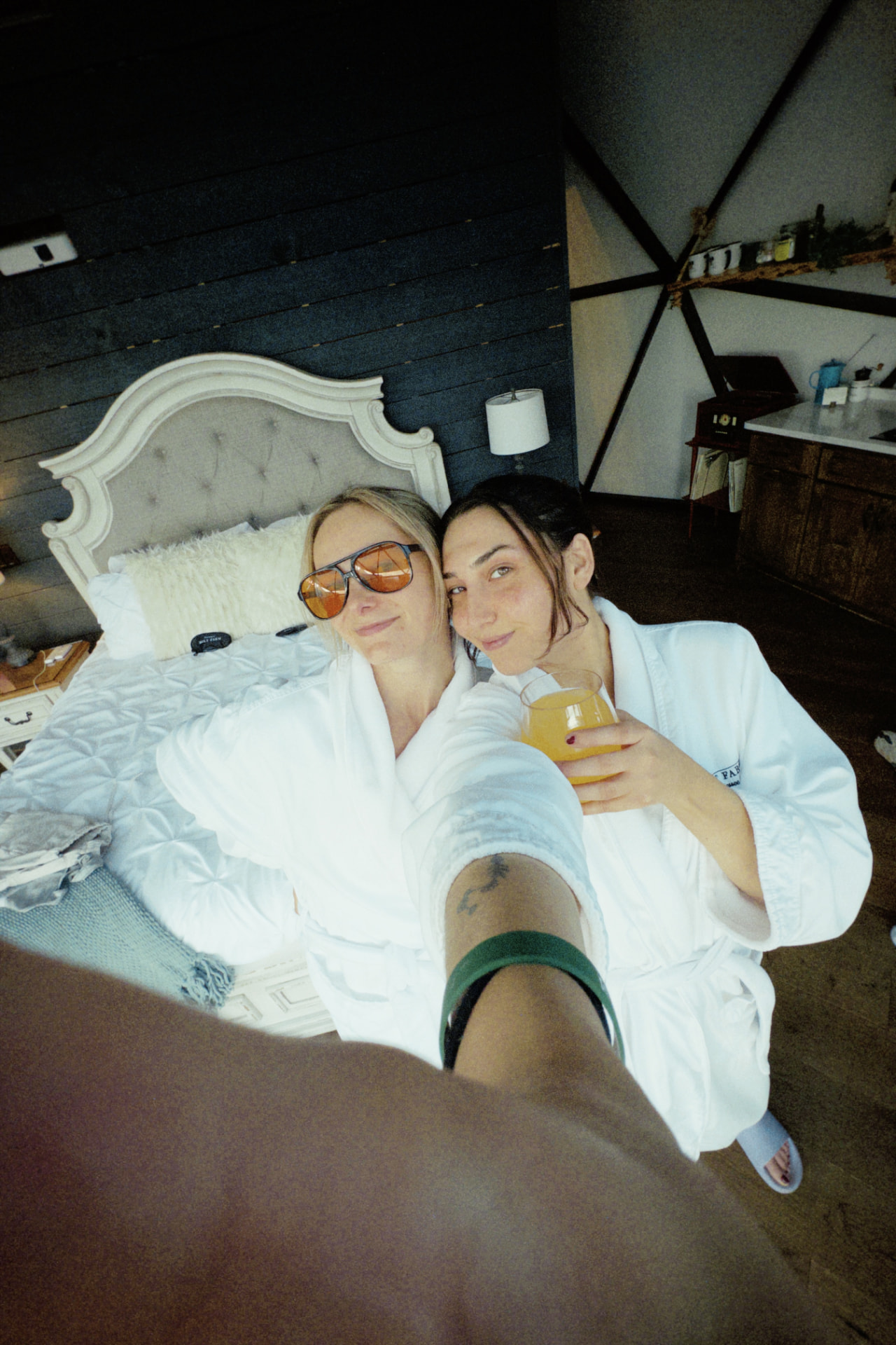 Two women in white robes leaning together on a bed, sharing a drink on a slow morning